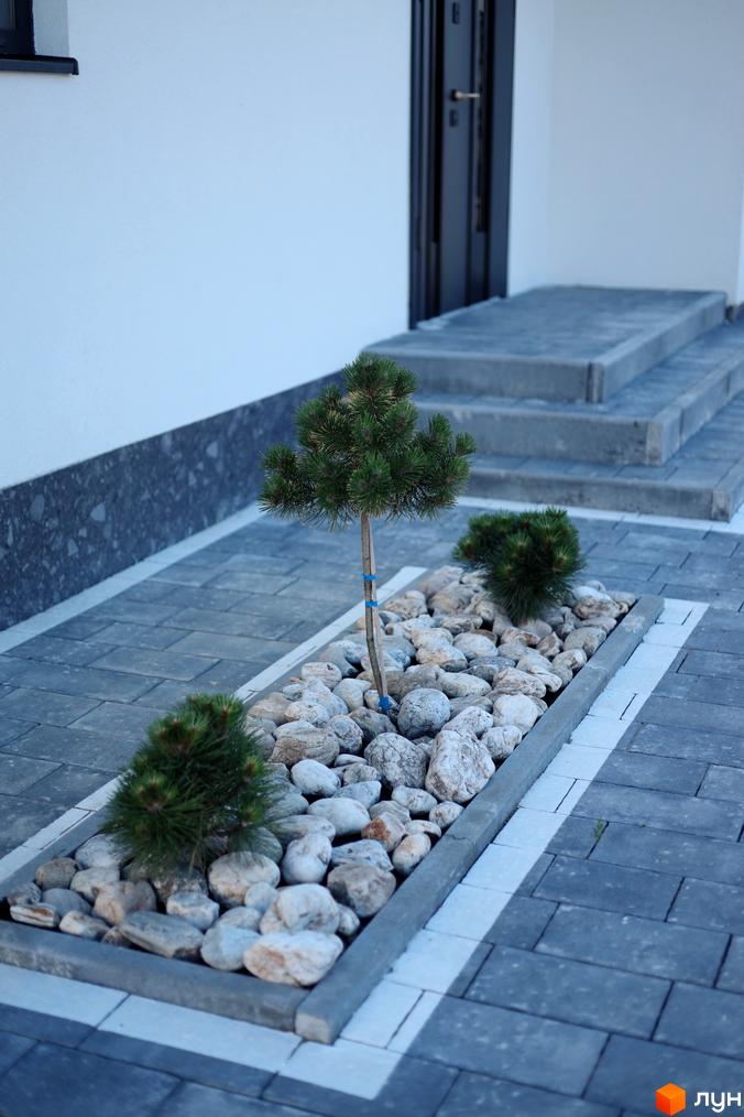 Фасад будинку з темними дверима та сходами. Прибудинкова територія з бруківкою, клумбою з камінням та декоративними деревами.