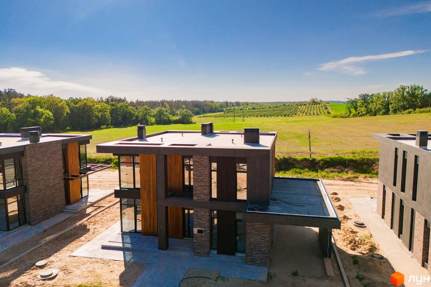 Будинки Котеджного містечка 9 ЕЛЕМЕНТІВ з темними фасадами, великими вікнами та оздобленням з дерева. Навколо ґрунтова ділянка, а на задньому плані зелені поля та ліс.