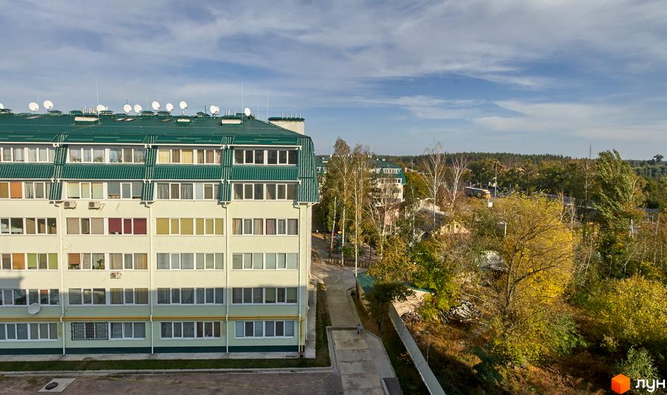 Зовнішній вигляд житлового комплексу Милі квартири з зеленим дахом та світлими фасадами. Навколо будинку розташована прибудинкова територія з деревами та доріжками.