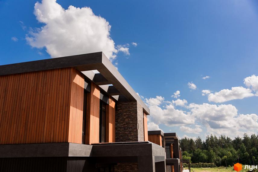 Зовнішній вигляд котеджного містечка КМ 9 ЕЛЕМЕНТІВ з дерев'яним фасадом та сірими елементами на фоні блакитного неба з хмарами.