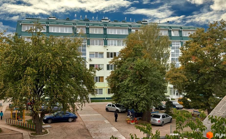 Зовнішній вигляд житлового комплексу Милі квартири з зеленим дахом та світлими стінами. Прибудинкова територія з деревами та припаркованими автомобілями.