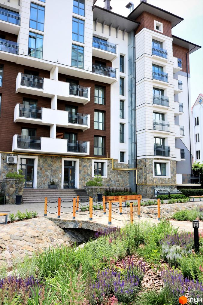 Новобудова Крайобраз з коричневими та білими фасадами, скляними балконами та кам'яною обробкою цоколя. Прибудинкова територія з доглянутими зеленими насадженнями та кам'яним мостом.