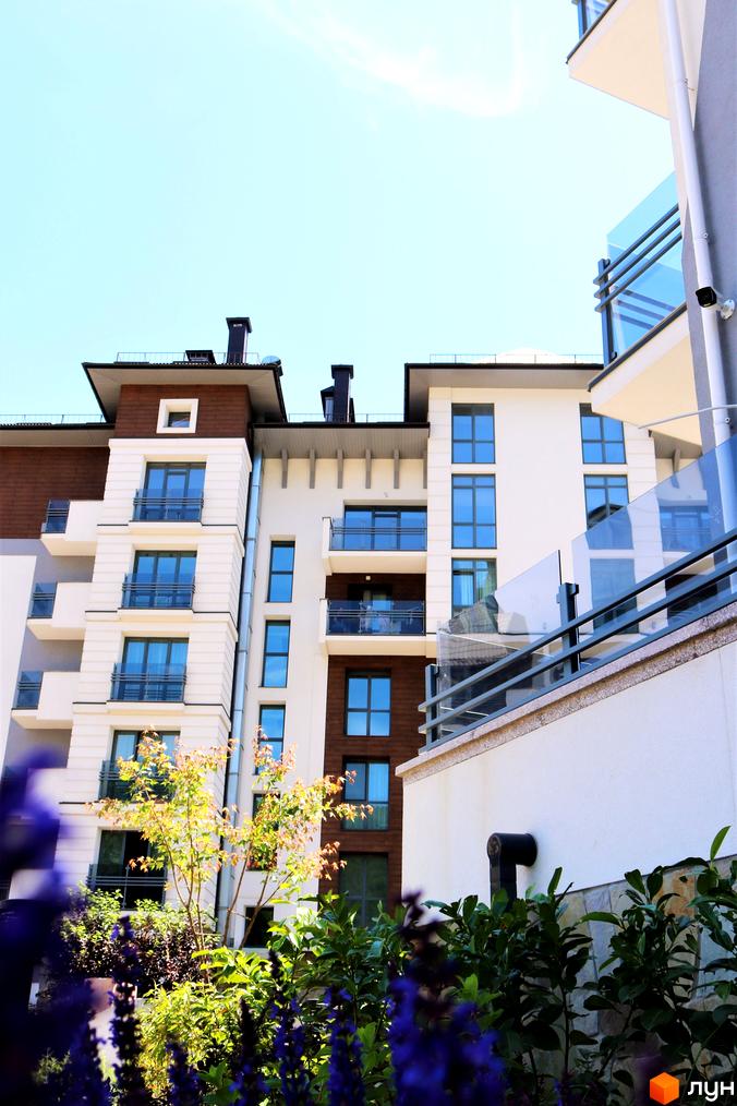 Новобудова Крайобраз з білими фасадами та коричневими вставками, скляними балконами та зеленими насадженнями на території. Сучасна архітектура житлового комплексу.