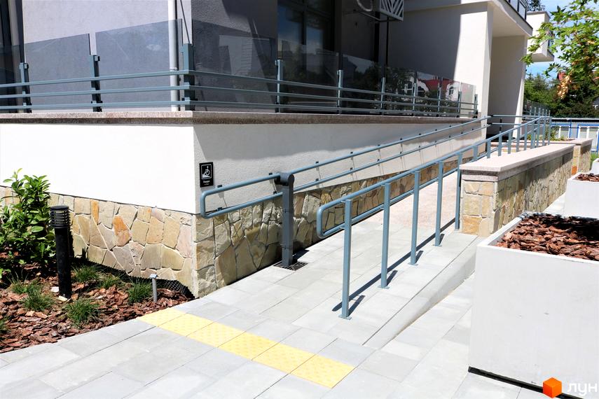 Зовнішній вигляд новобудови ЖК Крайобраз з пандусом для людей з інвалідністю та тактильною плиткою. Прибудинкова територія з озелененням та бетонними клумбами.