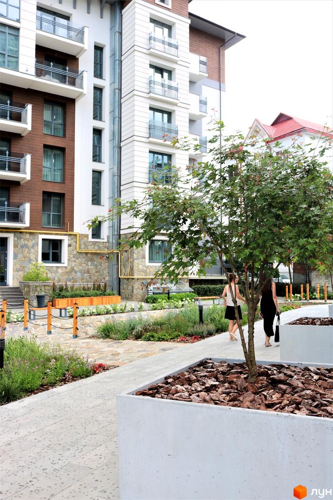 Житловий комплекс Крайобраз: фасади будівель з коричневими та білими елементами, балконами та кам'яною кладкою. Прибудинкова територія з доріжками, зеленню та деревами.