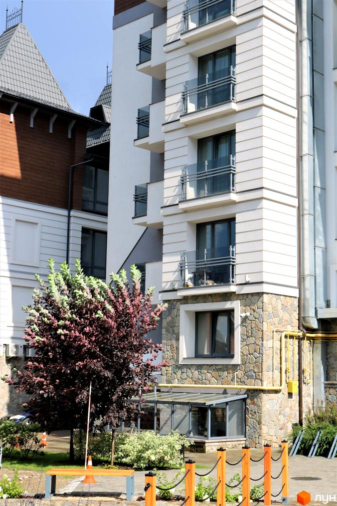 Сучасний житловий комплекс Крайобраз з кам'яним оздобленням цоколя та скляними балконами. На прибудинковій території розташовані зелені насадження та лавочка.