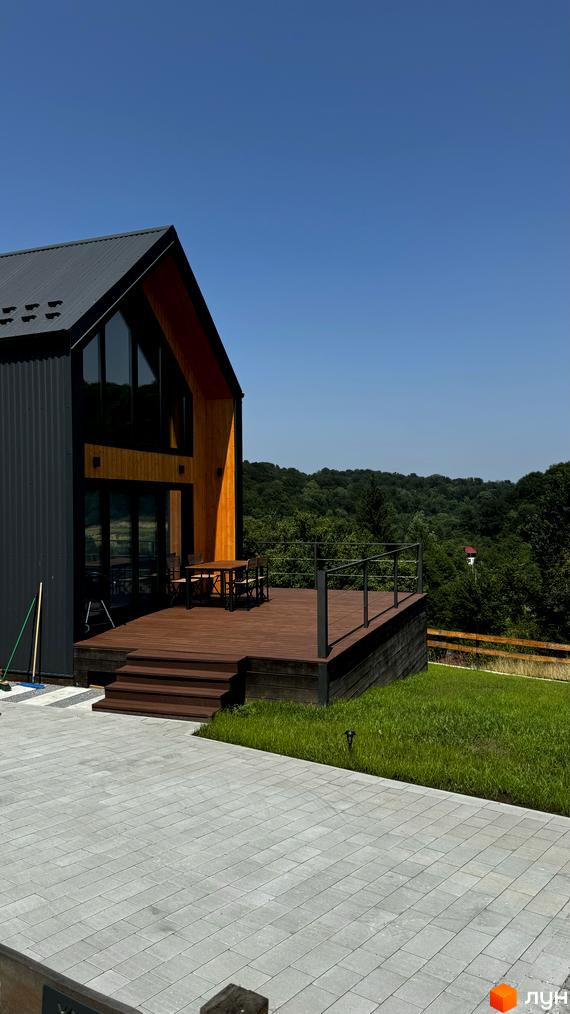 Сучасний котеджне містечко Схили з дерев'яною терасою та панорамними вікнами, оточений зеленню. Прибудинкова територія з бруківкою та доглянутим газоном.