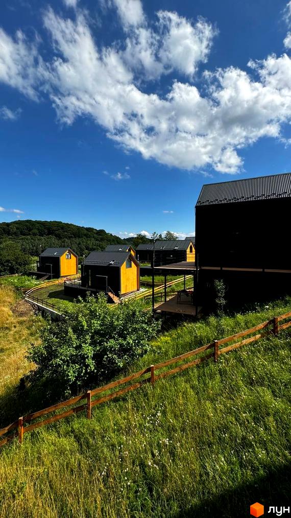 Котеджне містечко Схили: сучасні жовто-чорні будиночки на тлі зеленого пагорба та блакитного неба з хмарами. Територія огороджена дерев'яним парканом.