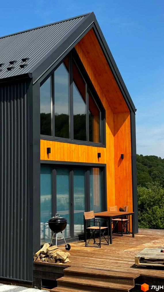 Котеджне містечко КМ Схили: двоповерховий будинок з панорамними вікнами та дерев'яним оздобленням, тераса з барбекю та місцем для відпочинку.