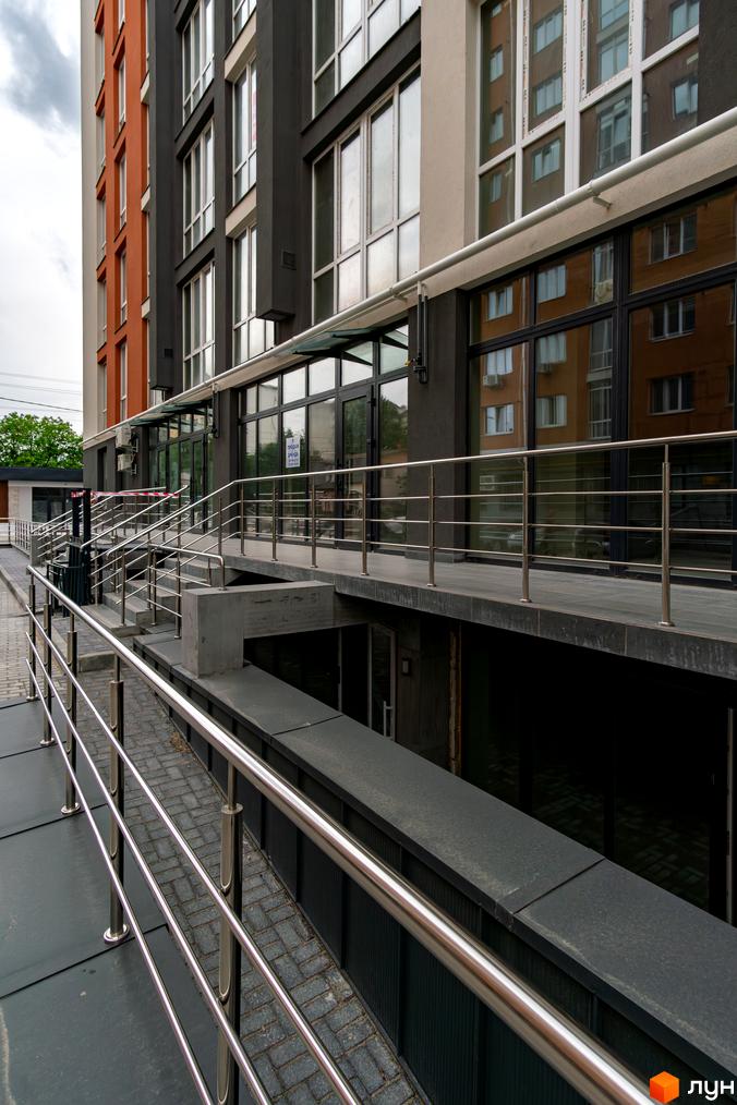 Зовнішній вигляд новобудови ЖК Затишний з цегляним фасадом та великими вікнами. Поруч розташовані пандус та сходи з металевими поручнями.