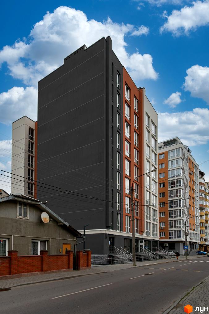 Зовнішній вигляд новобудови ЖК Затишний з фасадами темного та цегляного кольору. Поруч розташована цегляна огорожа та дорога.