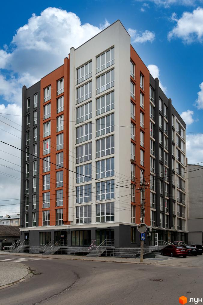 Зовнішній вигляд житлового комплексу Затишний з фасадами сірого, помаранчевого та білого кольорів. Навколо будинку облаштована прибудинкова територія з доріжками та місцями для паркування.