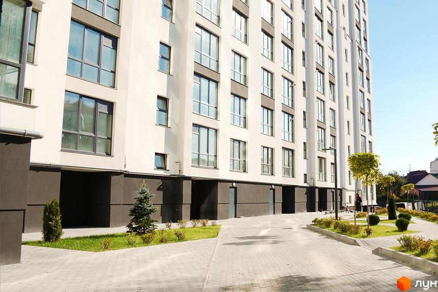 Житловий комплекс Затишний: багатоповерховий будинок світлого кольору з великими вікнами та облаштована прибудинкова територія з доріжками та зеленню.