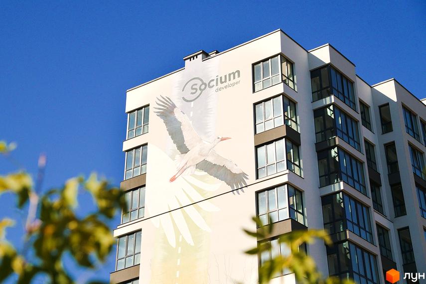 Новобудова Затишний: багатоповерховий будинок світлого кольору з великим малюнком лелеки на фасаді. На передньому плані видно зелені гілки.