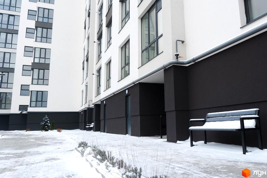 Фасад Житлового комплексу Затишний у біло-темних кольорах з великими вікнами. Засніжена прибудинкова територія з лавкою та рослинами.