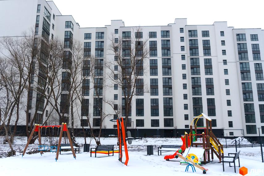 Висока будівля ЖК Затишний з великою кількістю вікон. На передньому плані дитячий майданчик, вкритий снігом.
