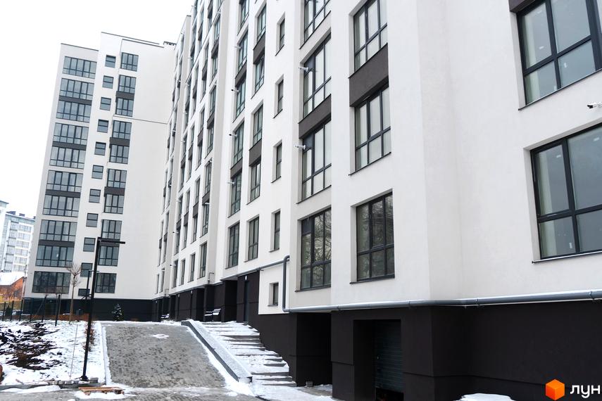 Білосніжний багатоповерховий будинок ЖК Затишний з великими вікнами. На території є сходи, пандус та доріжки, вкриті снігом.