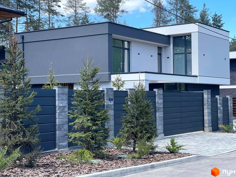 Котеджне містечко КМ Нова Ходосівка Premium – фасад будинку з великими вікнами та сірою обшивкою, огороджений парканом з бетонних блоків та темних панелей.