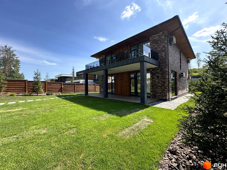 Котеджне містечко КМ Нова Ходосівка Premium. Двоповерховий будинок з цегляним фасадом, скляним балконом та зеленим газоном перед ним.