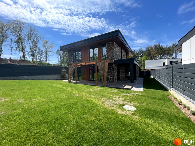 Котеджне містечко Нова Ходосівка Premium. Зовнішній вигляд будинку з кам'яним фасадом та дерев'яними елементами, зелена галявина та паркан.