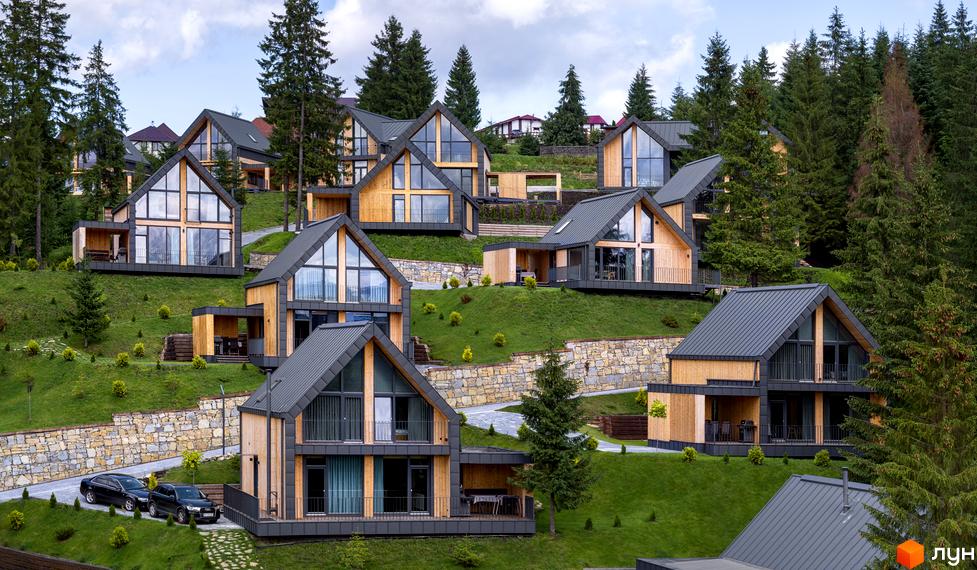 Зображення Котеджного містечка SKOGUR з дерев'яними котеджами на зеленій галявині серед сосен. На передньому плані припарковані автомобілі.