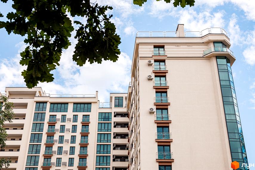 Новобудова HydroPark DeLuxe з бежевими фасадами, балконами зі скляними огорожами та кутовим склінням. На передньому плані зелені дерева.