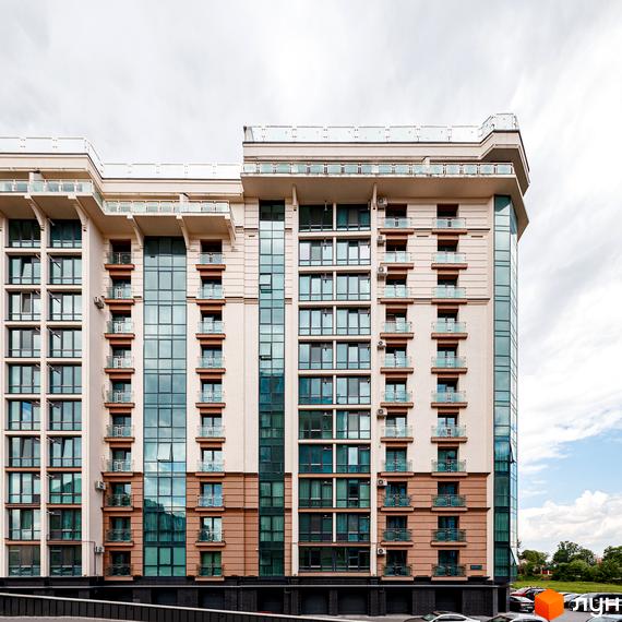 Житловий комплекс HydroPark DeLuxe: багатоповерховий будинок світлого кольору з великими вікнами та балконами. На передньому плані видно парковку, а на задньому – зелені насадження.