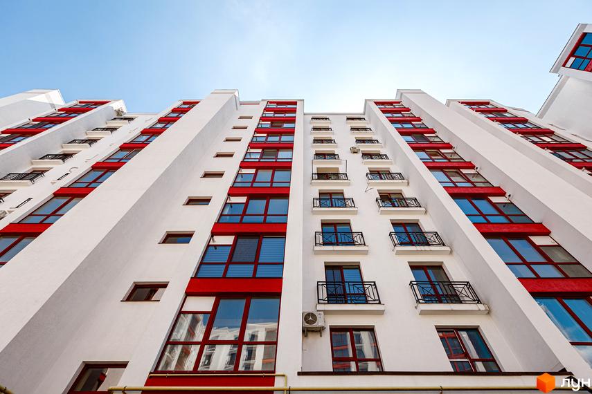 Висотна новобудова з білими фасадами та червоними акцентами. На балконах ковані огорожі, навколо будівлі блакитне небо.