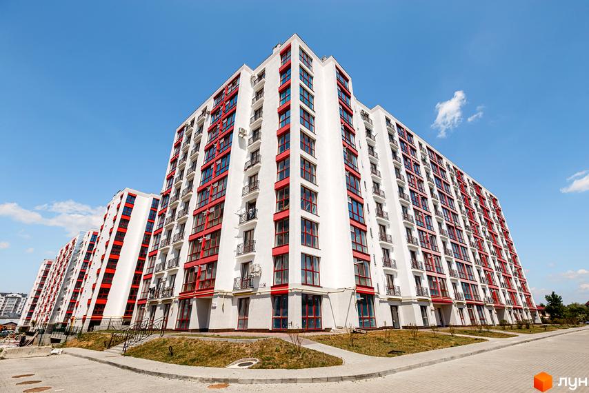 Білі фасади з червоними віконними рамами та балконами житлового комплексу Містечко Козацьке. Перед будинками прокладена дорога та зелені насадження.