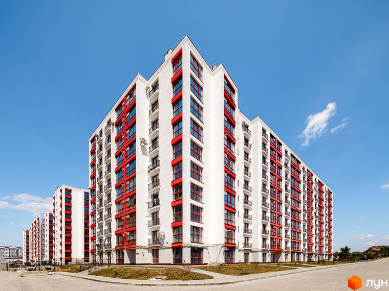 Білосніжні багатоповерхівки з яскраво-червоними акцентами на фасадах. На передньому плані — асфальтована територія та зелені насадження.