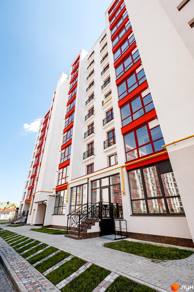 Висотна будівля житлового комплексу з червоними акцентами на фасаді. На передньому плані доглянута прибудинкова територія з пішохідними доріжками та газонами.