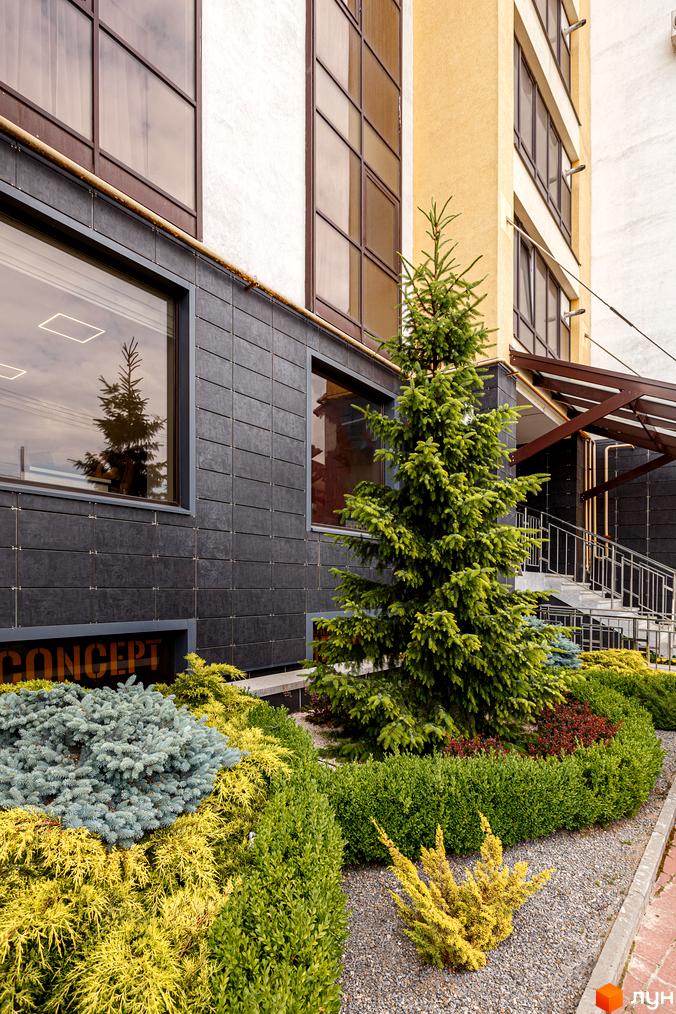 Фасад Житловий комплекс Lystopad оздоблений темною плиткою та має великі вікна. Навколо висаджені декоративні хвойні дерева та кущі.