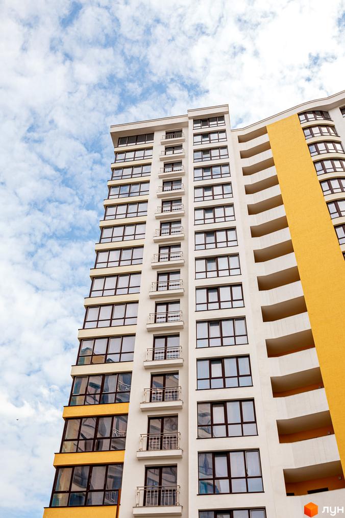 Зовнішній вигляд житлового комплексу Lystopad з білими та жовтими фасадами, великими вікнами та балконами на тлі блакитного неба з хмарами.