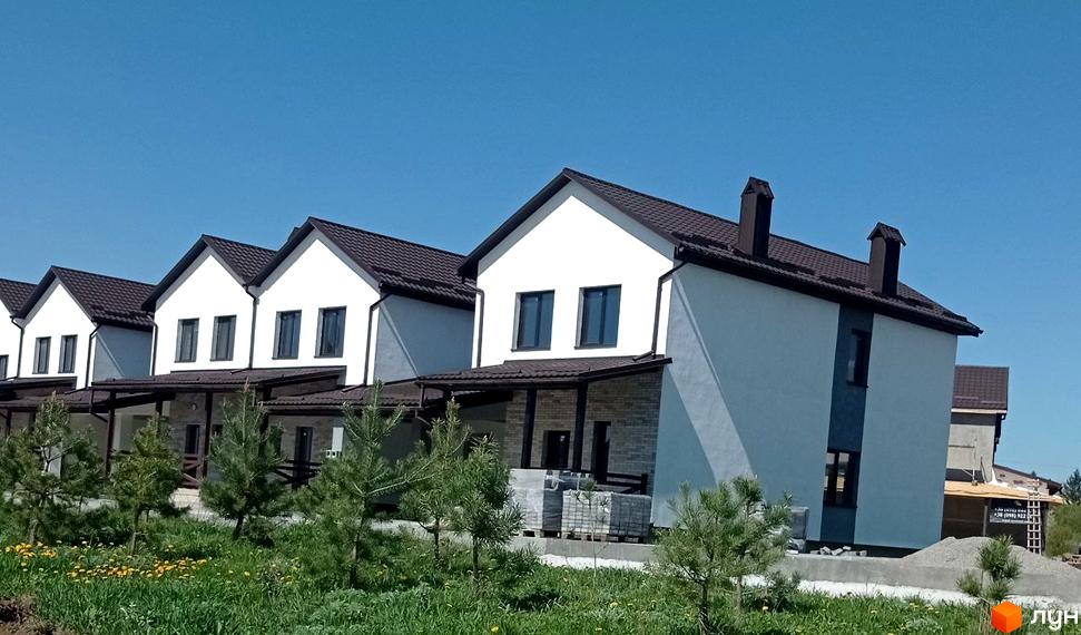 Котеджне містечко Затишний пропонує білі двоповерхові будинки з коричневими дахами та зеленими насадженнями навколо. На території комплексу розташовані молоді сосни та газон із жовтими квітами.