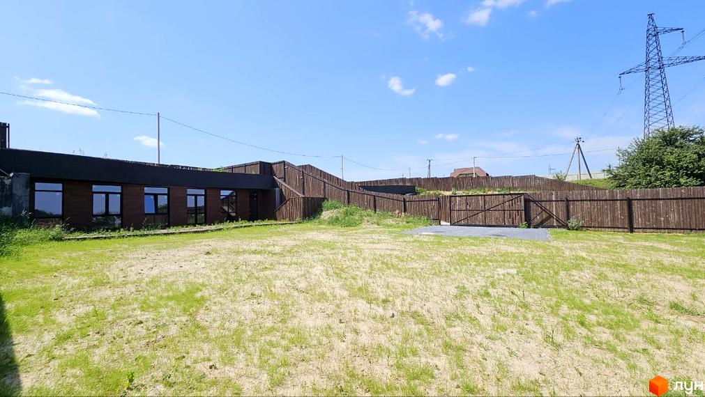 Котеджне містечко KRAIEVYD: одноповерхова будівля з темними вікнами та дерев'яним фасадом, огороджена дерев'яним парканом. Прибудинкова територія з газоном та воротами.