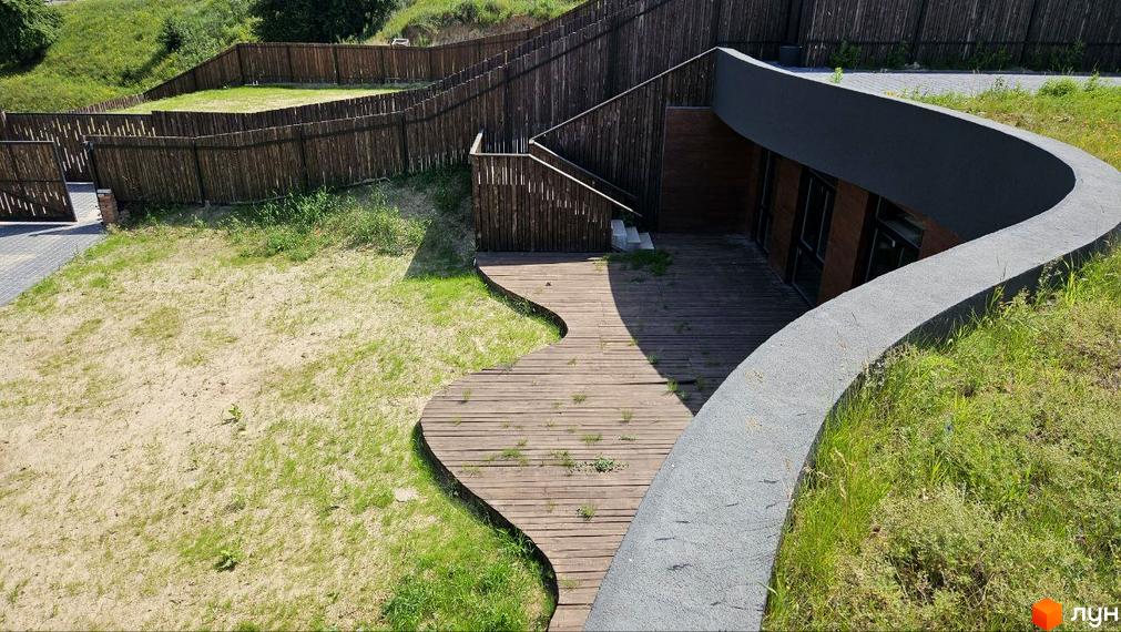 Котеджне містечко KRAIEVYD: будинок з вигнутою сірою стіною та дерев'яною терасою. Прибудинкова територія з парканом та зеленими насадженнями.