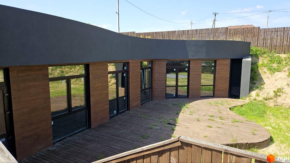 Котеджне містечко KRAIEVYD. Зовнішній вигляд будинку з дерев'яною обшивкою та великими вікнами. Тераса з дерев'яним покриттям та зеленню навколо.