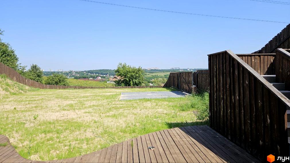 Котеджне містечко KRAIEVYD: вид на зелену галявину з дерев'яними сходами та парканом. На задньому плані видно панораму міста та пагорби.