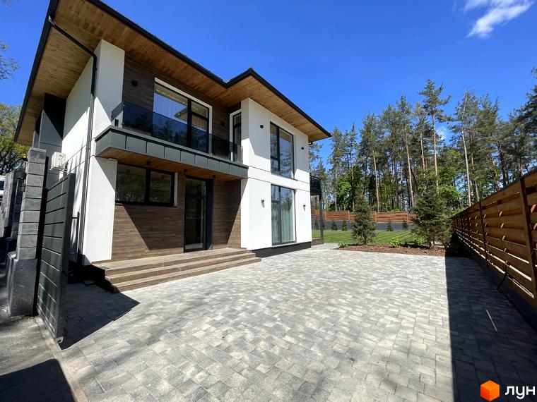 Котеджне містечко Нова Ходосівка: двоповерховий будинок з білим та дерев'яним фасадом, великими вікнами та балконом. Перед будинком брукований двір та зелена територія з деревами.