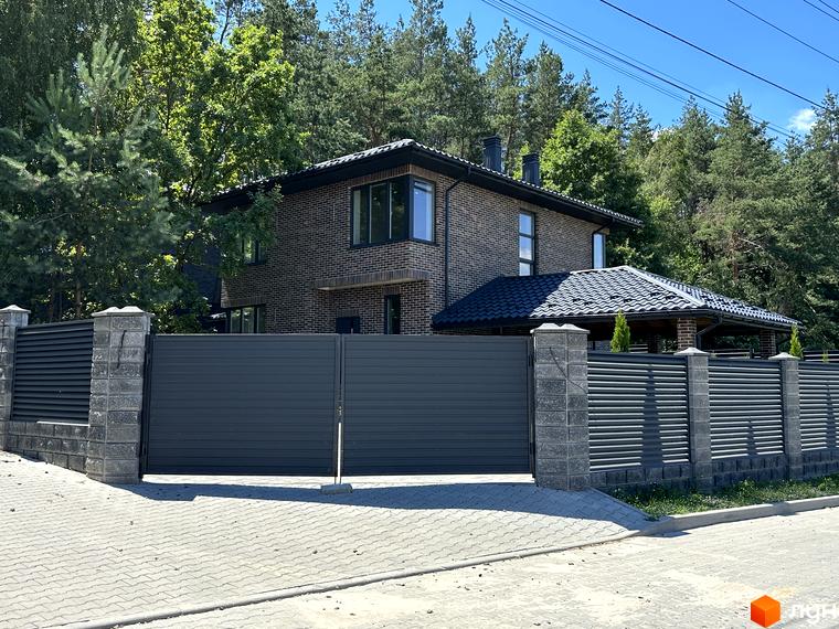 Котеджне містечко Нова Ходосівка: цегляний будинок з темним дахом, паркан з кам'яними стовпами та воротами, серед дерев.