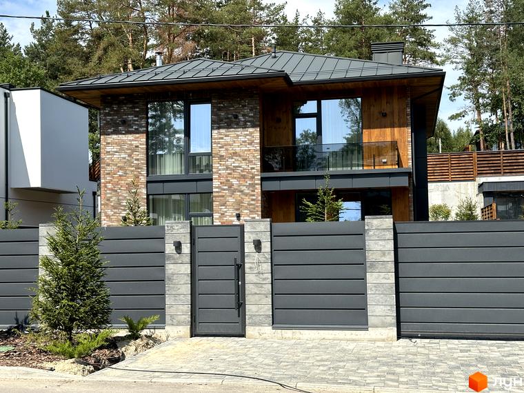 Двоповерховий будинок Котеджного містечка Нова Ходосівка з цегляним та дерев'яним оздобленням, великими вікнами. Перед будинком сірий паркан та хвойні дерева.