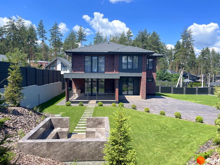 Двоповерховий будинок Котеджного містечка Нова Ходосівка з темно-червоної цегли та темними вікнами. Перед будинком зелений газон, вимощена доріжка та ландшафтний дизайн.