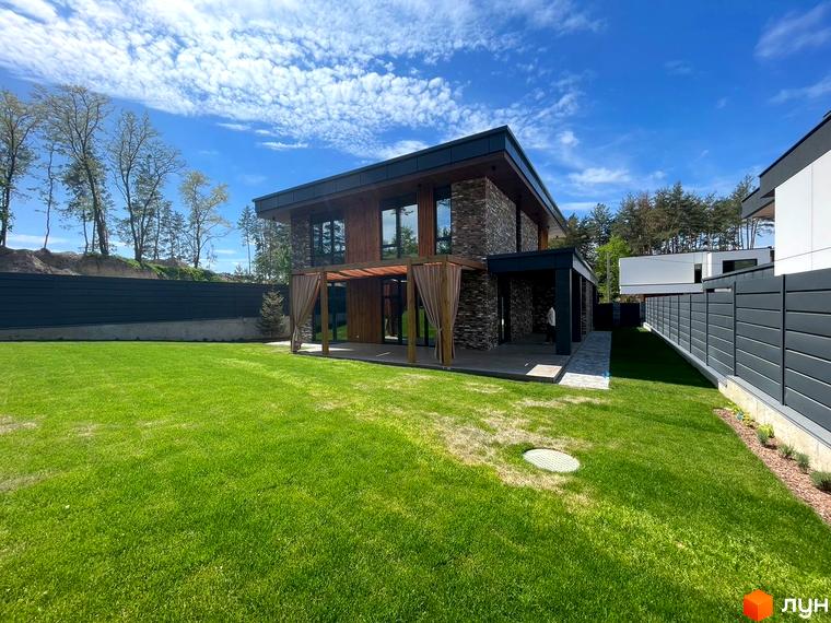 Двоповерховий будинок Котеджного містечка Нова Ходосівка з темним фасадом та великими вікнами. Перед будинком зелений газон та тераса з перголою.