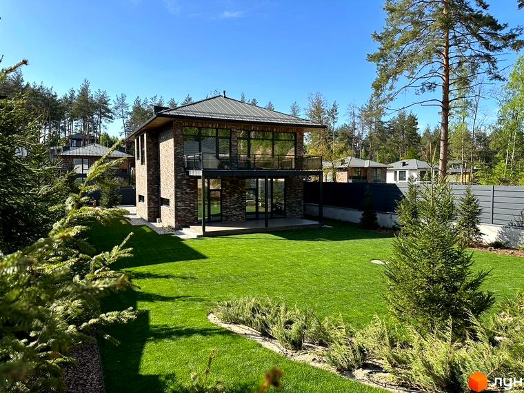 Котеджне містечко Нова Ходосівка: двоповерховий будинок з цегляним фасадом, панорамними вікнами та балконом. Навколо доглянутий газон та хвойні дерева.