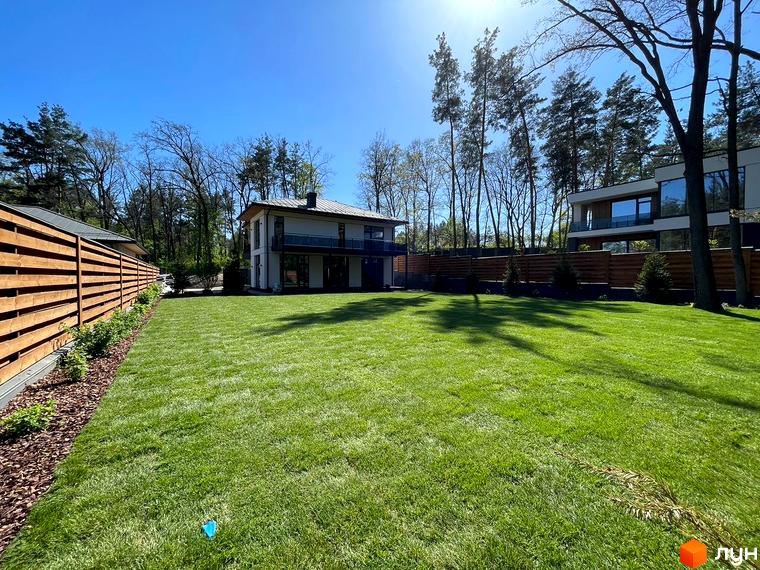 Зображення будинку в Котеджному містечку Нова Ходосівка з великою галявиною перед ним. На задньому плані дерева та інші будинки.