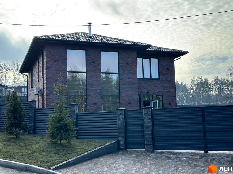 Зображення двоповерхового будинку з червоної цегли в Котеджному містечку Нова Ходосівка. Навколо будинку зелений газон та дерева.