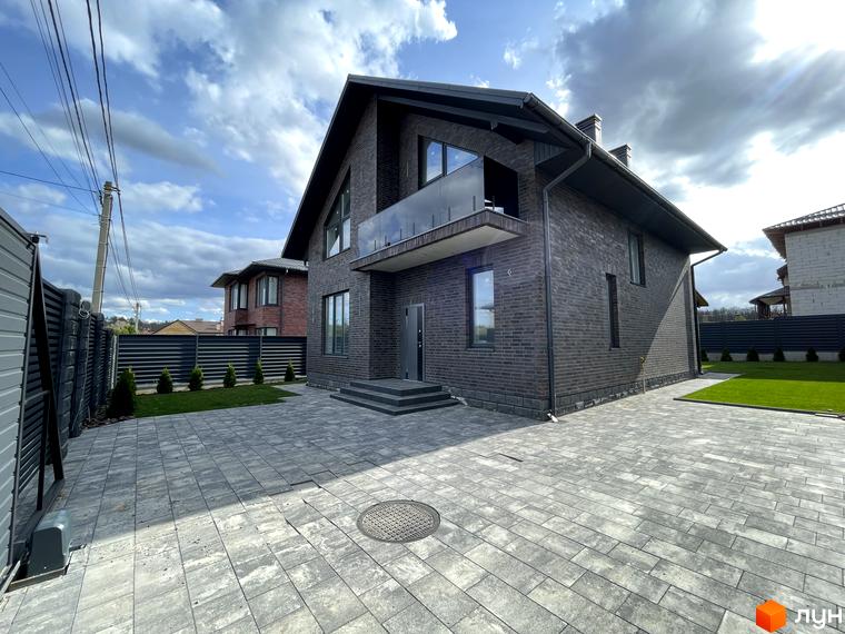 Темний цегляний будинок з великими вікнами та балконом у Котеджному комплексі Нова Ходосівка. Прибудинкова територія з бруківкою та зеленим газоном.