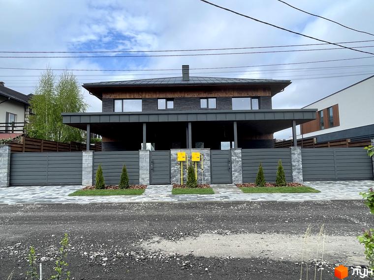 Котеджне містечко Нова Ходосівка: двоповерховий будинок з темним дахом та фасадом, великі вікна. Перед будинком сірий паркан, брукована доріжка та зелені насадження.