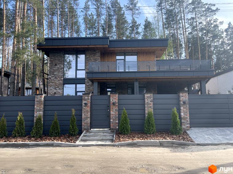 Котеджне містечко Нова Ходосівка: двоповерховий будинок з цегляним фасадом, темними вікнами та балконом. Перед ним сірий паркан з туями.