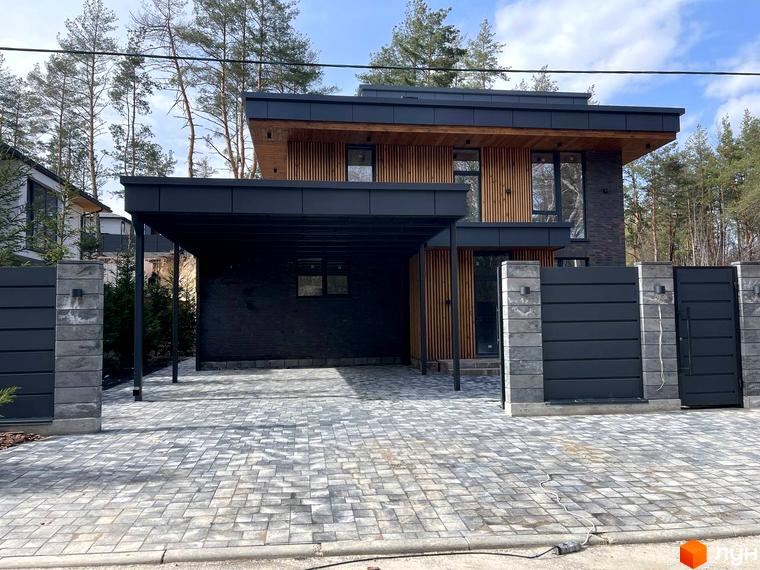 Зовнішній вигляд двоповерхового будинку в Котеджному містечку Нова Ходосівка з чорним дахом та дерев'яним оздобленням. Подвір'я викладене бруківкою.